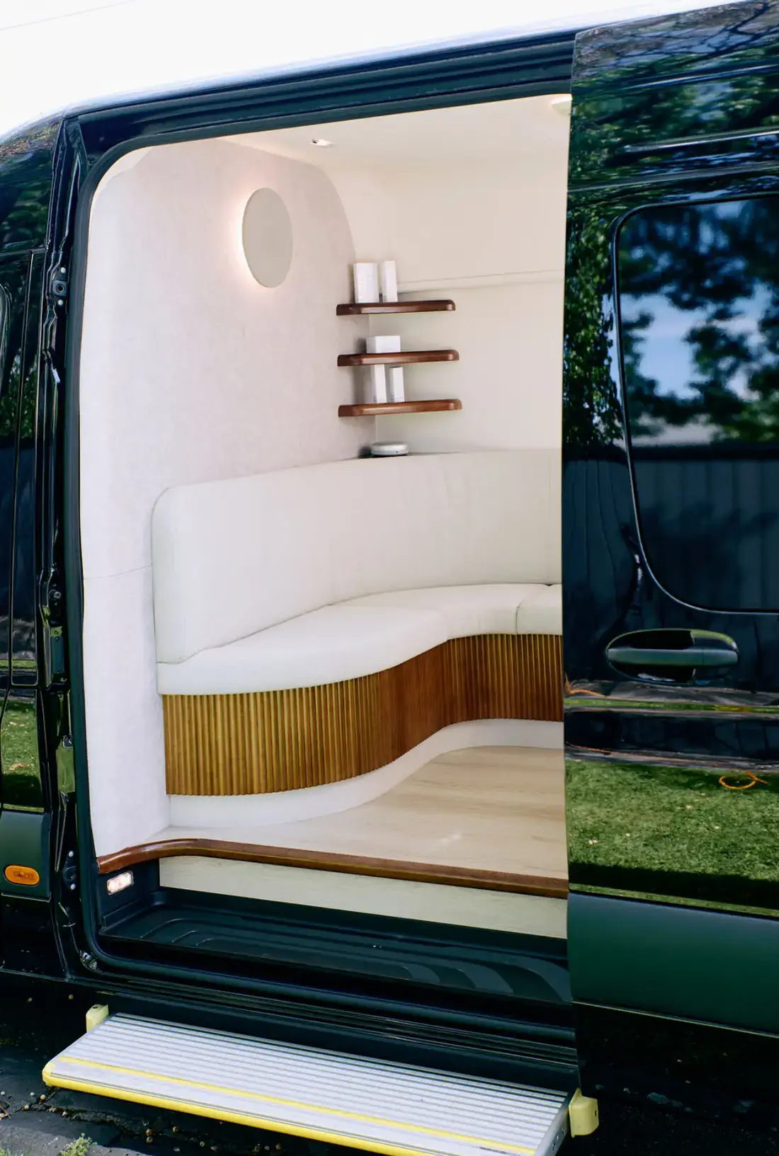 Through the open side door of the Sprinter, a curved upholstered banquette on a fluted timber base, with rounded shelves and a circular illuminated mirror