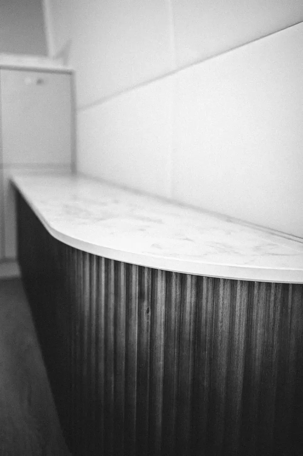 Black and white detail of a curved sink cabinet faced in fluted timber, under a stone-look worktop