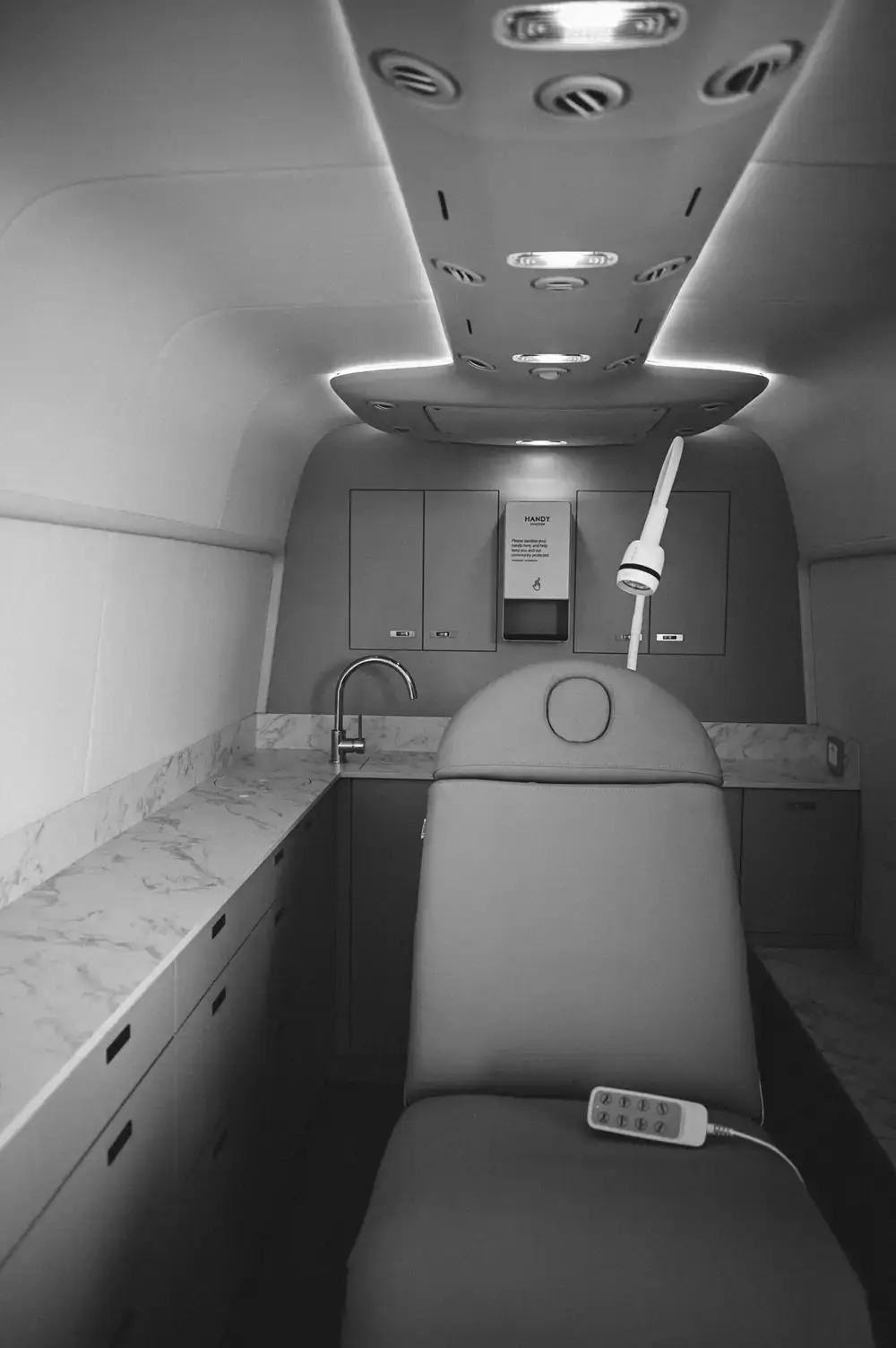 Black and white view along the treatment area: the treatment bed, task light, stone-look work surfaces and rear cabinetry under a lit ceiling