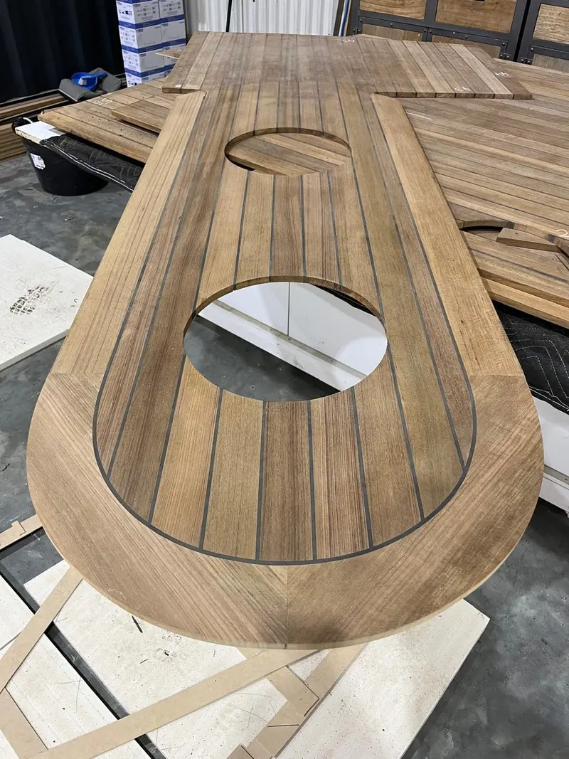 A shaped teak deck panel with a curved margin and two round cut-outs, laid on the workshop floor