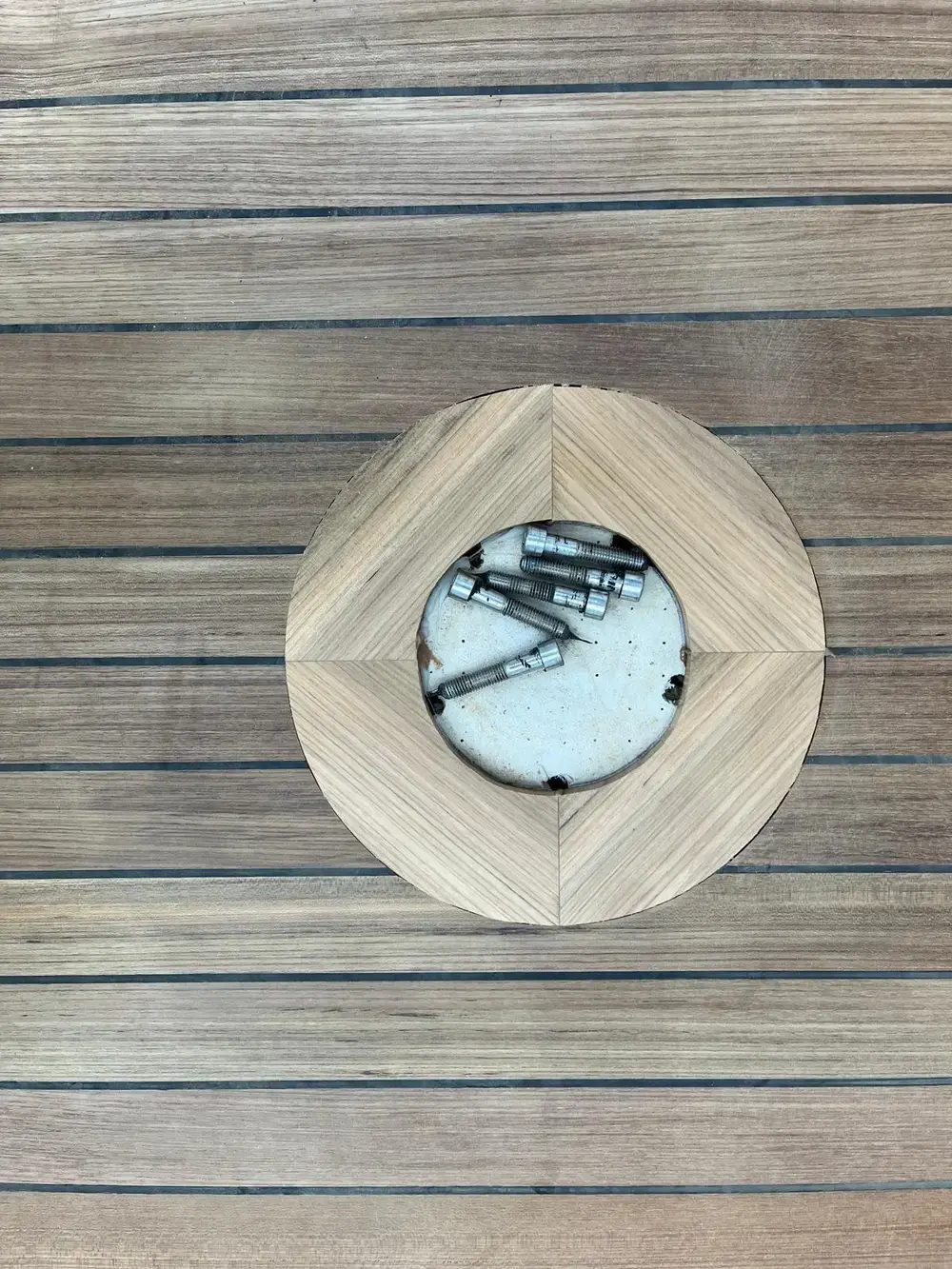 A weathered teak plank field with dark seams around a round deck hatch framed by shaped margin pieces