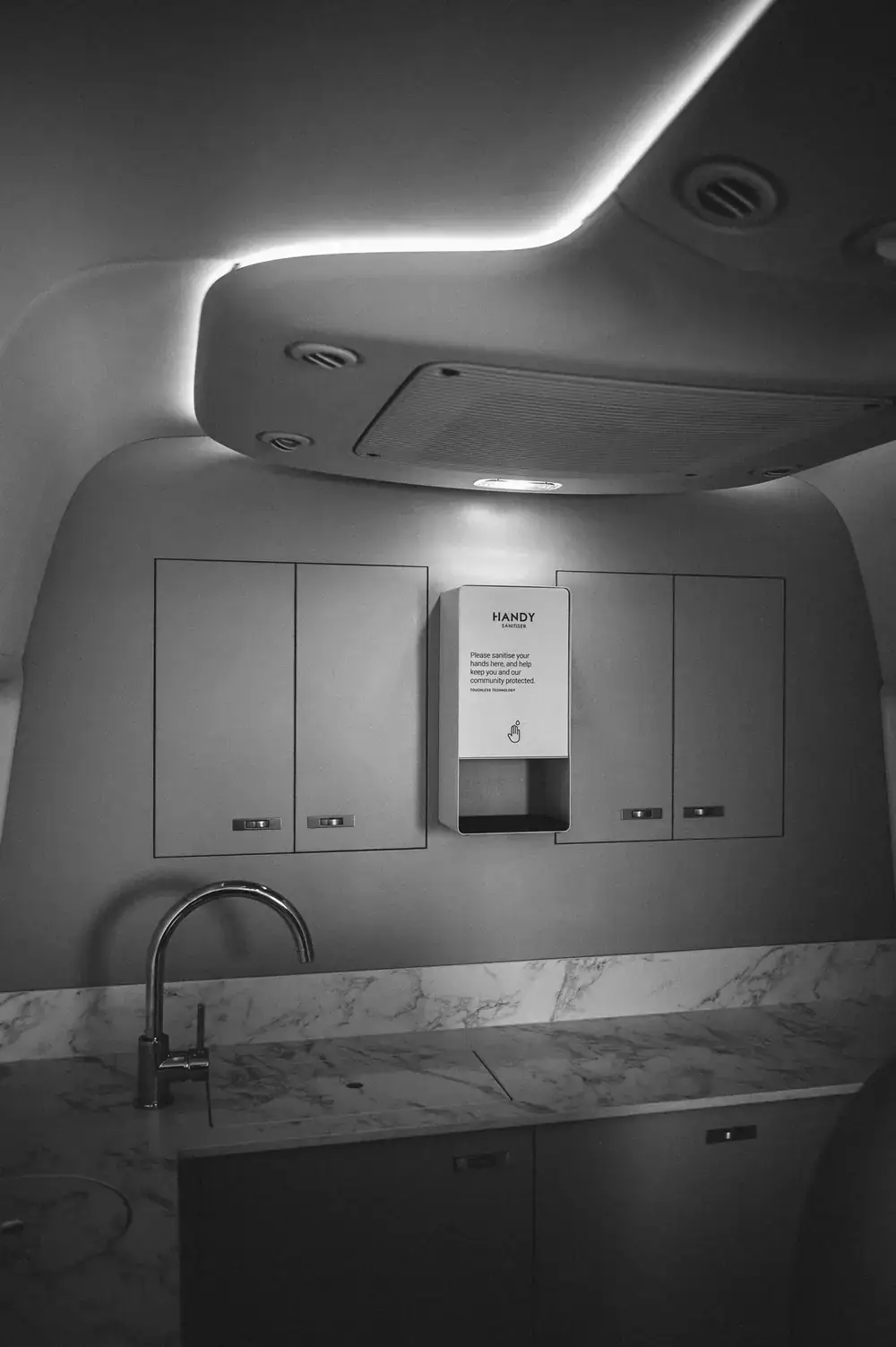 Black and white view of flush wall cabinets with a touchless sanitiser, above a stone-look worktop and tap