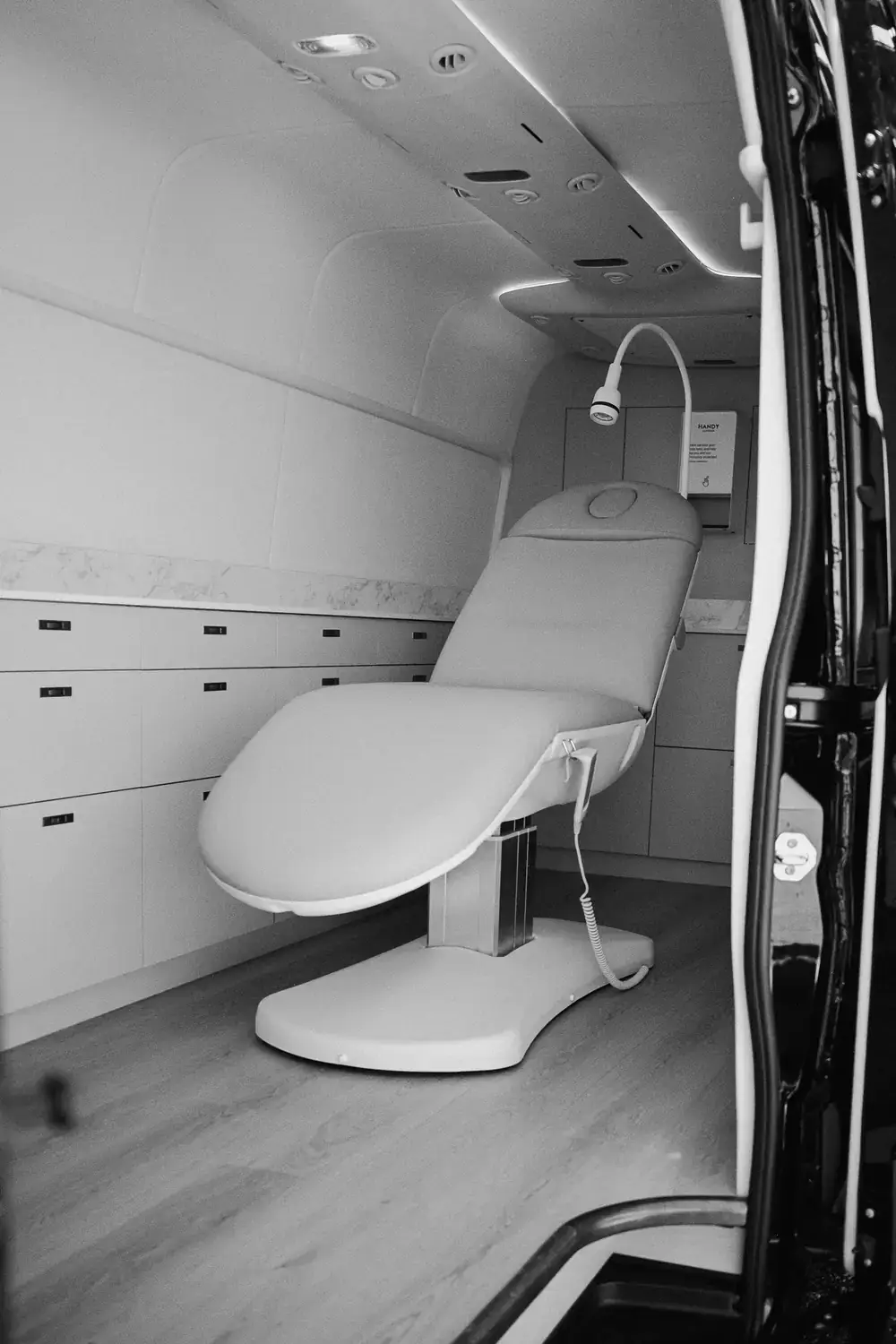 Black and white view through the rear door of the Sprinter: the treatment bed on a timber-look floor, with a bank of drawers along the wall