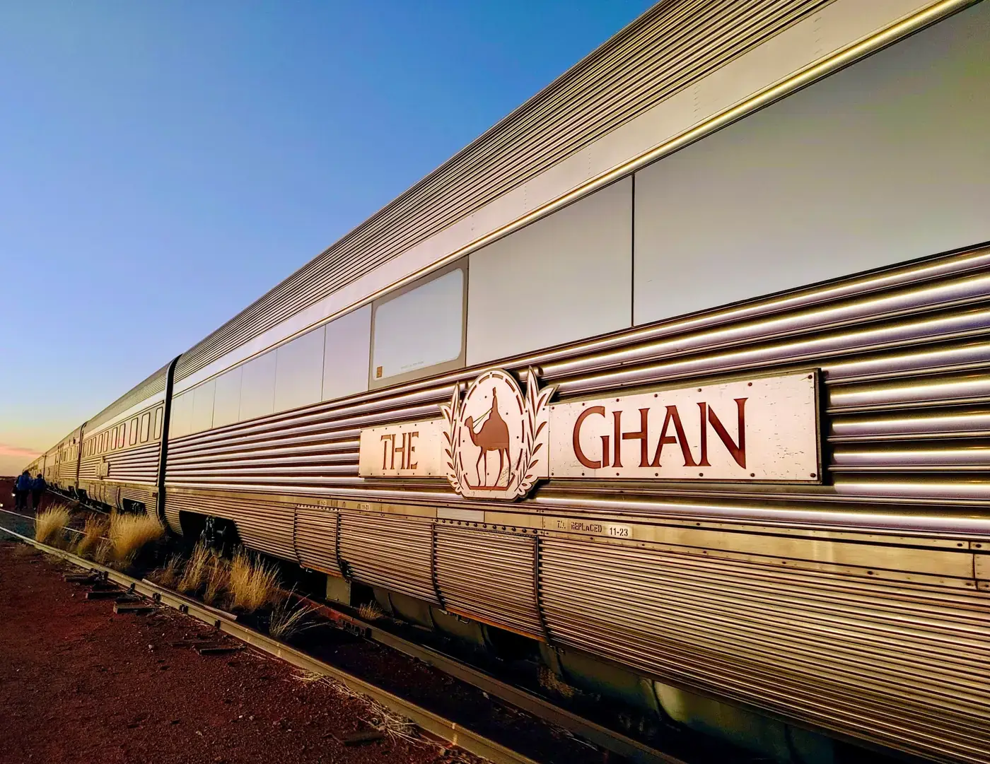 The stainless steel carriages of The Ghan, with its camel emblem, standing on red earth under a clear dusk sky